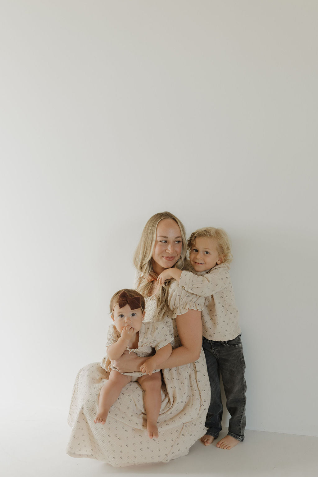 A woman sits on the floor with a baby in her lap and a young child hugging her, all wearing coordinated forever french baby Women's Short Sleeve Muslin Dress | Flora in light hues.