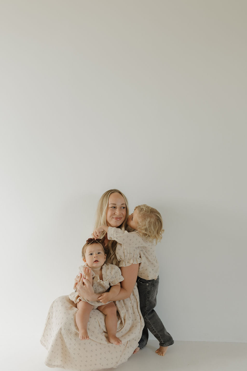A woman smiles against a white wall as a young child kisses her cheek and a baby sits on her lap. All wear matching forever french baby Women's Short Sleeve Muslin Dress | Flora in soft neutrals. The background is simple and uncluttered.