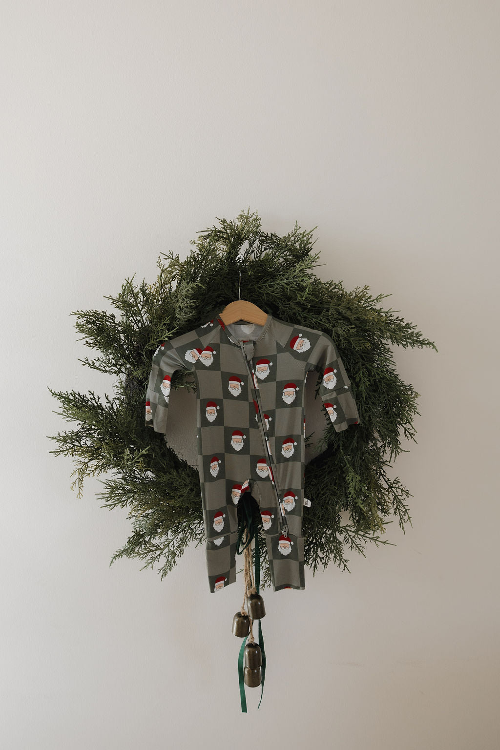 A green wreath hangs on a white wall displaying forever french baby Bamboo Zip Pajamas | Santa Checkerboard in the center, with a cluster of bells tied with green ribbons hanging below.