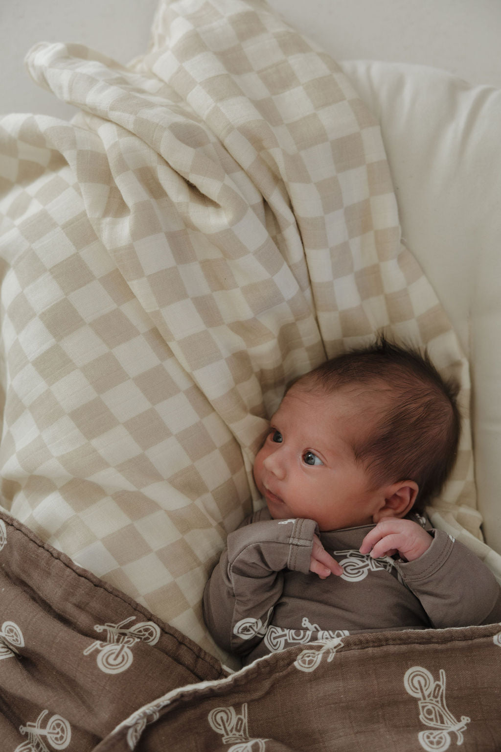 A newborn lies on a bed, wrapped in the forever french baby Muslin Quilt | Dirt Bike & Checkerboard and resting on a beige checkered pillow, gazing up with wide eyes.