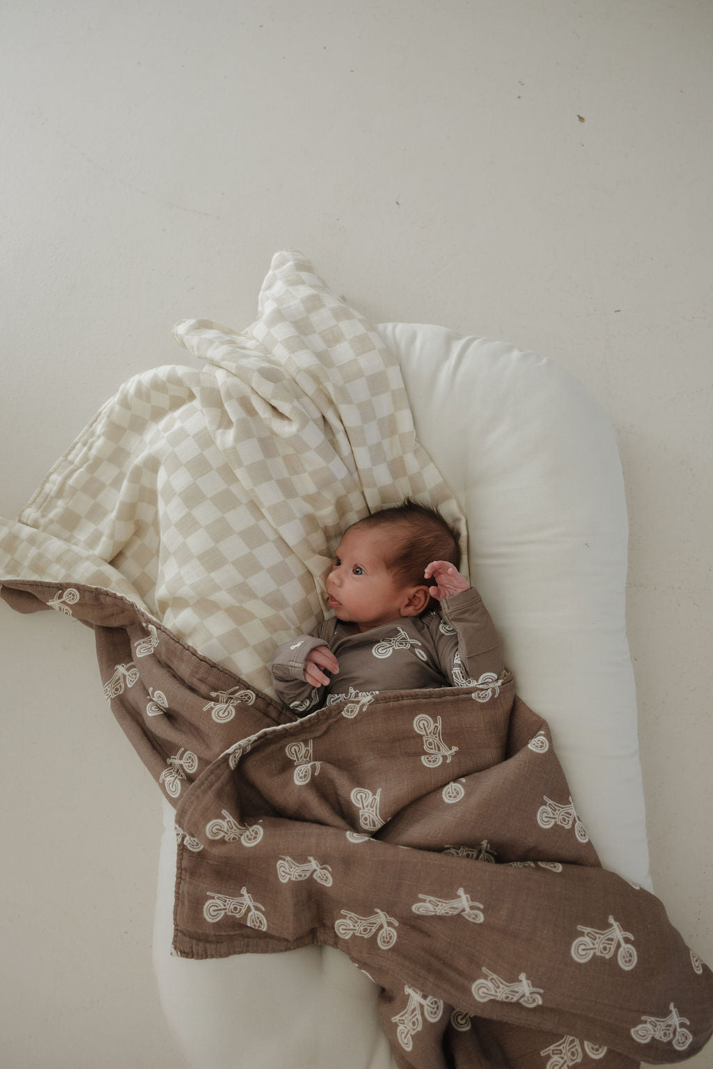A baby lies on a white cushioned surface, wrapped in the forever french baby Muslin Quilt | Dirt Bike & Checkerboard, gazing upward with one hand near their head.