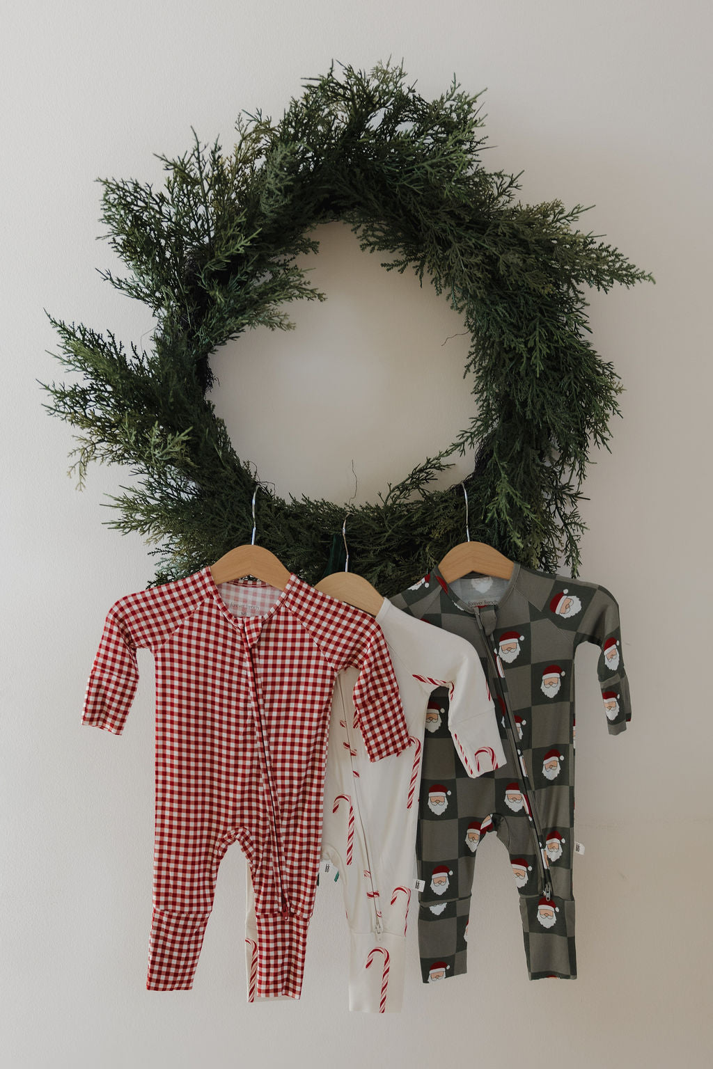 A large green wreath hangs on a white wall with three baby onesies—including the forever french baby Bamboo Zip Pajamas in Red Gingham—all crafted from breathable sleepwear for your little one's comfort.