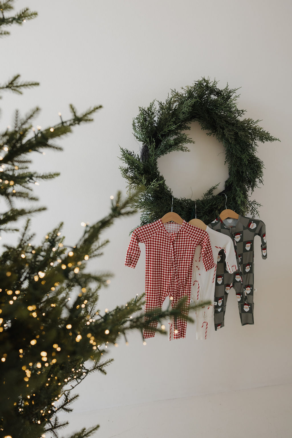 A Christmas tree glows with twinkling lights in front of a green wreath and three forever french baby Bamboo Zip Pajamas—Red Gingham, white, and green—soft, breathable, and festive sleepwear for little ones in holiday patterns.