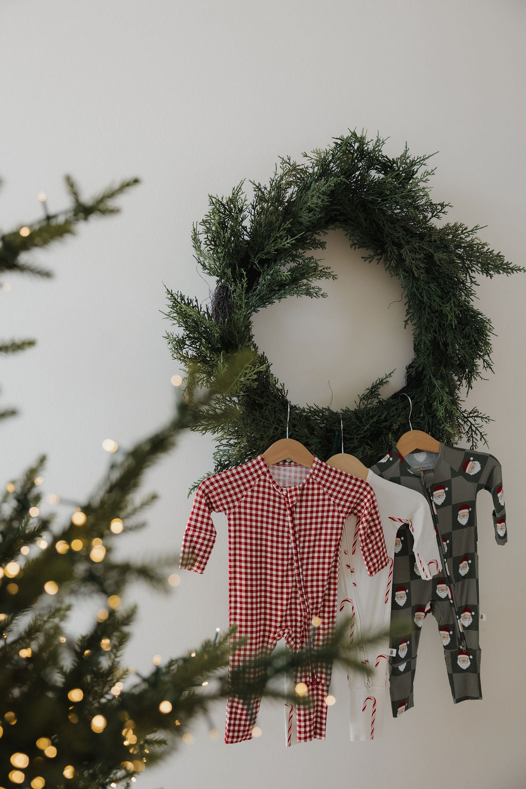 A green wreath hangs on a white wall with three forever french baby Bamboo Zip Pajamas—Santa Checkerboard, white with candy canes, and green with mugs. A blurred Christmas tree with lights is seen in the foreground.