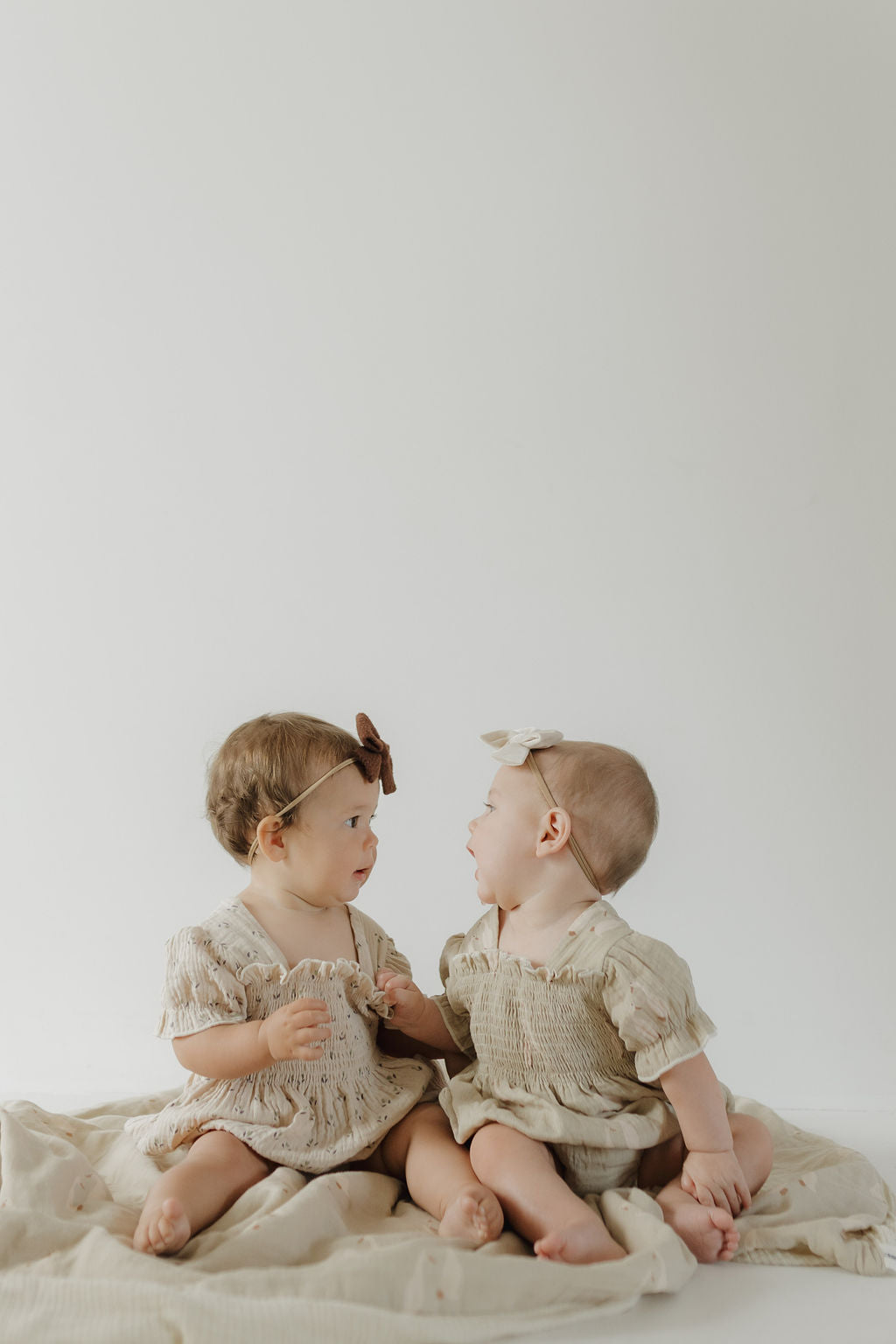 Two babies sit on the forever french baby Muslin Quilt | Silly Goose, wearing neutral outfits. One wears a brown headband, the other a white bow. They face each other and interact in this charming nursery moment against a plain background.