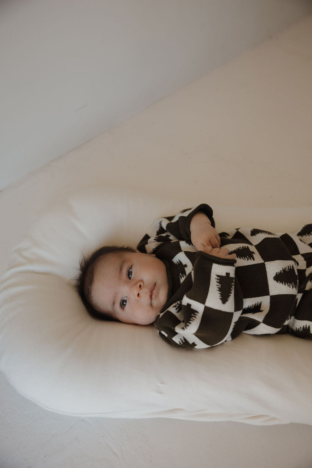 A baby rests on a soft white cushion, wearing the forever french baby Knit Pant Set | Christmas Tree Checkerboard—cozy 100% cotton with black and white geometric tree patterns—ideal for any winter wardrobe.