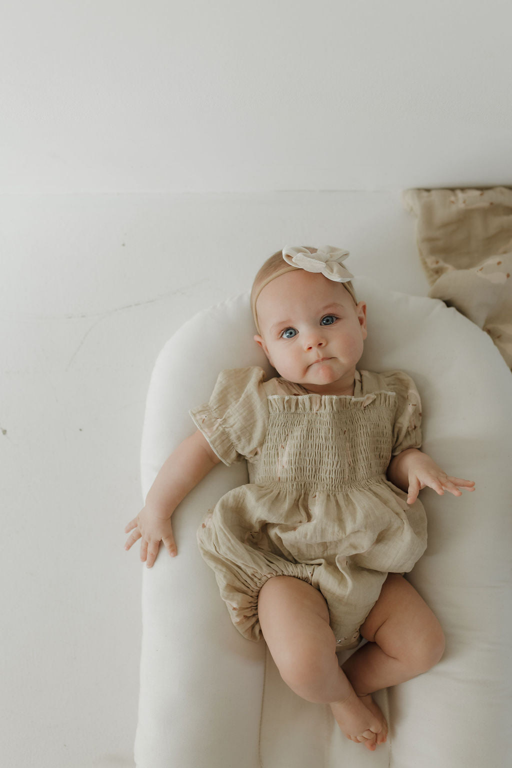 A baby with blue eyes, wearing the forever french baby Ruffle Muslin Romper | Silly Goose and a matching bow headband, lies calmly on a white cushion looking up.