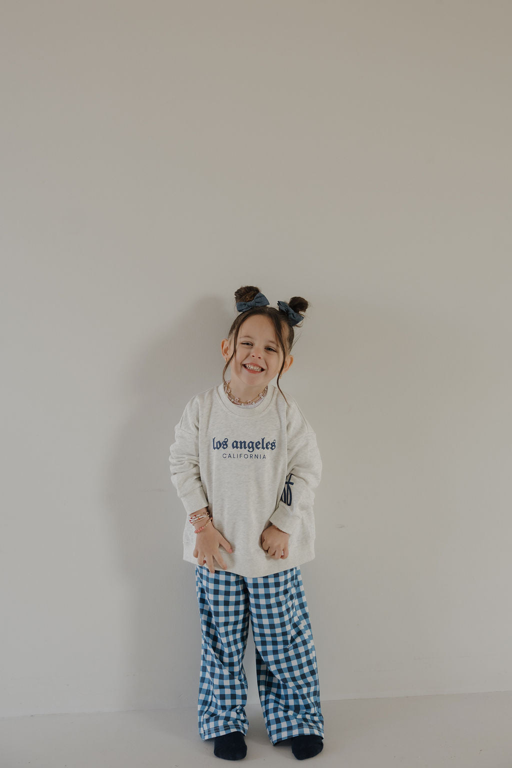 A young child with two buns, wearing the forever french baby Child Sweatshirt | ff Los Angeles and blue plaid pants, smiles against a plain light-colored wall.