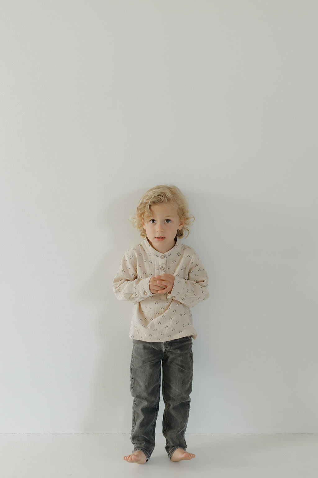 A young child with curly blonde hair stands barefoot against a white wall, wearing forever french baby’s Muslin Button Up | Flora and dark gray pants—a charming kids' look—with hands clasped, looking forward.