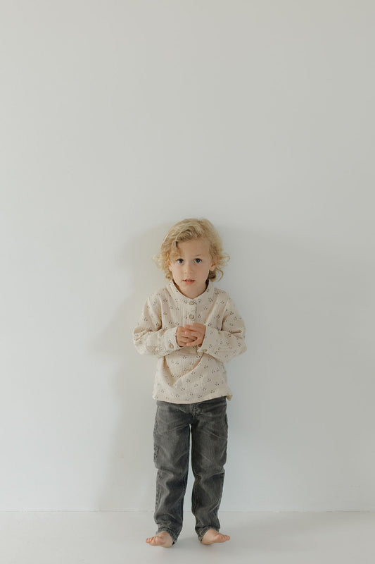 A young child with curly blonde hair stands barefoot against a white wall, wearing forever french baby’s Muslin Button Up | Flora and dark gray pants—a charming kids' look—with hands clasped, looking forward.