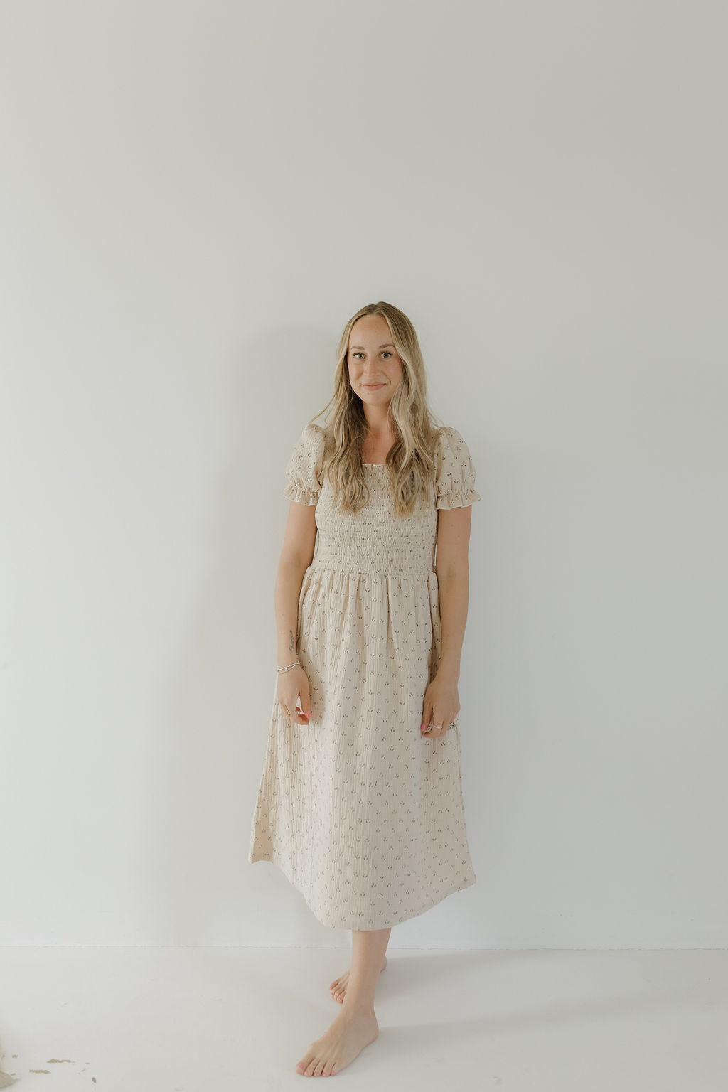 A woman with long blonde hair stands barefoot against a white wall, wearing the forever french baby Women's Short Sleeve Muslin Dress | Flora—lightweight and subtly patterned, perfect for mom-and-me matching.