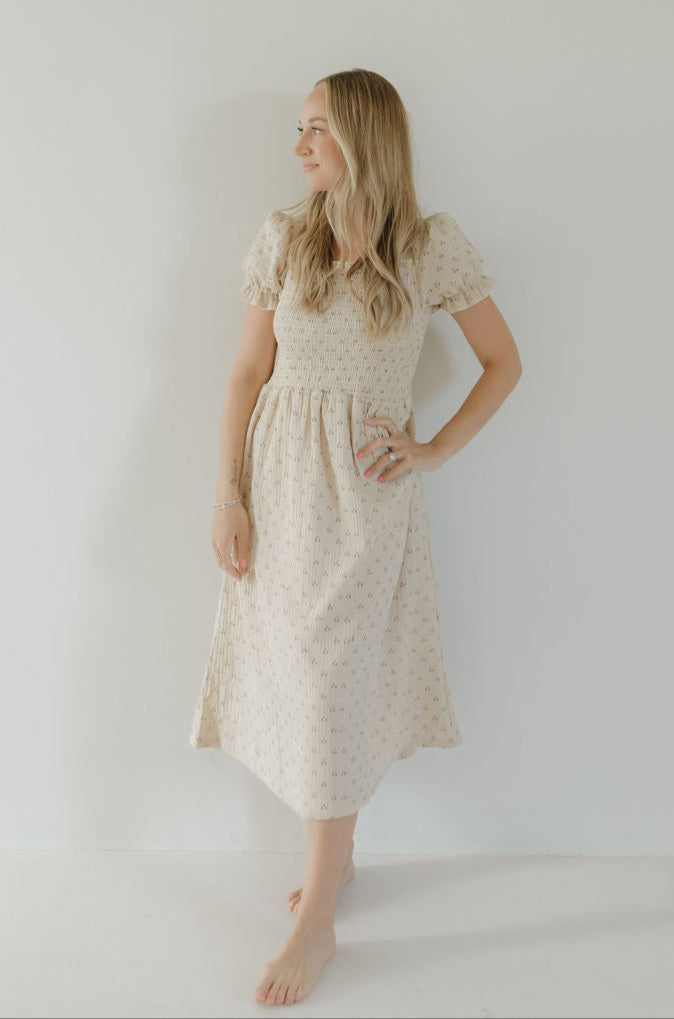A woman with long blonde hair stands barefoot against a white wall, wearing the forever french baby Women's Short Sleeve Muslin Dress | Flora, featuring short sleeves and a knee-length hem, looking left with one hand on her hip.