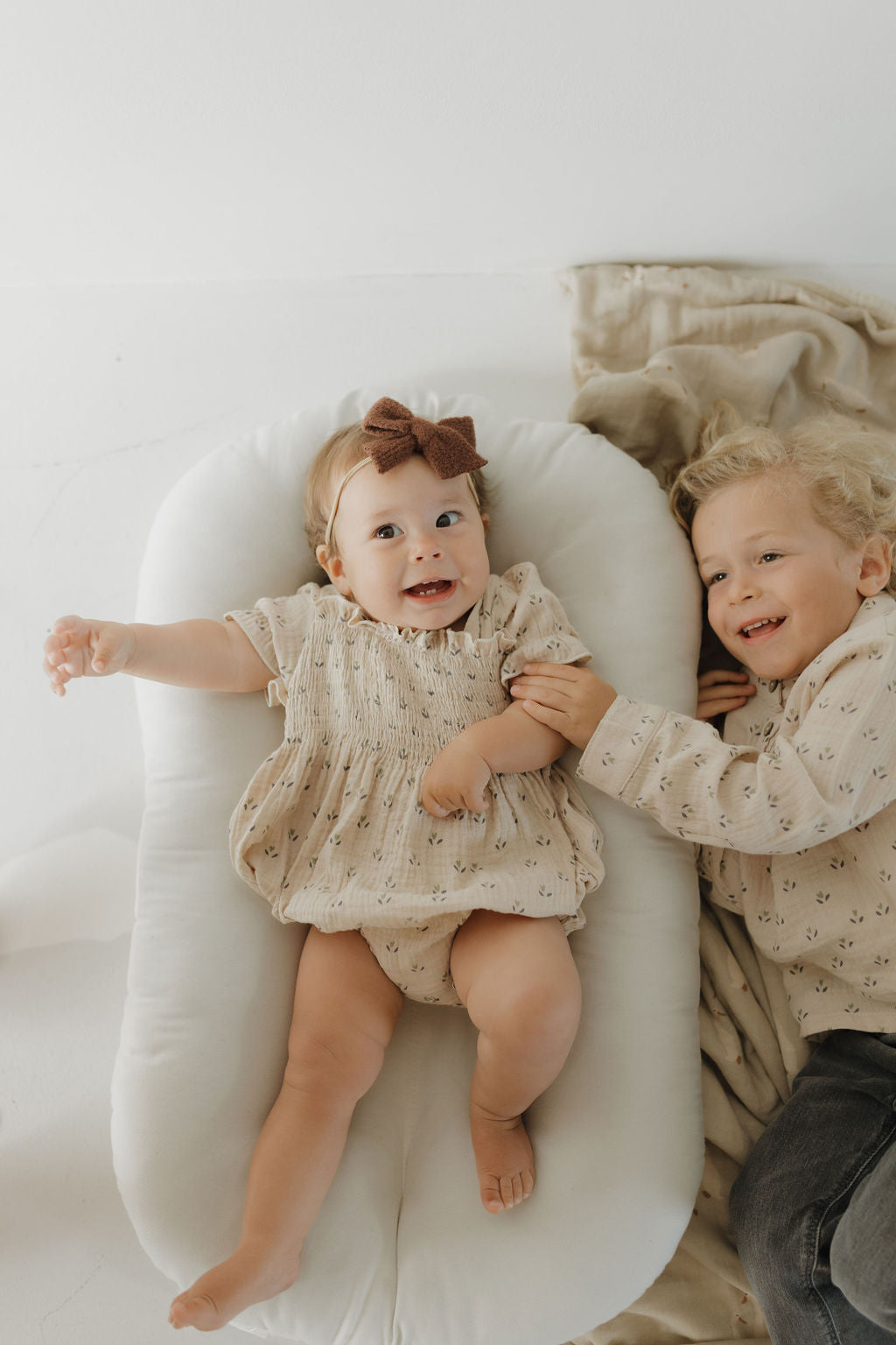 A baby in a beige muslin cotton dress and brown bow lies smiling on a cushion. Next to her, a young child wears the Muslin Button Up | Flora by forever french baby and smiles while holding the baby's hand on a soft blanket.