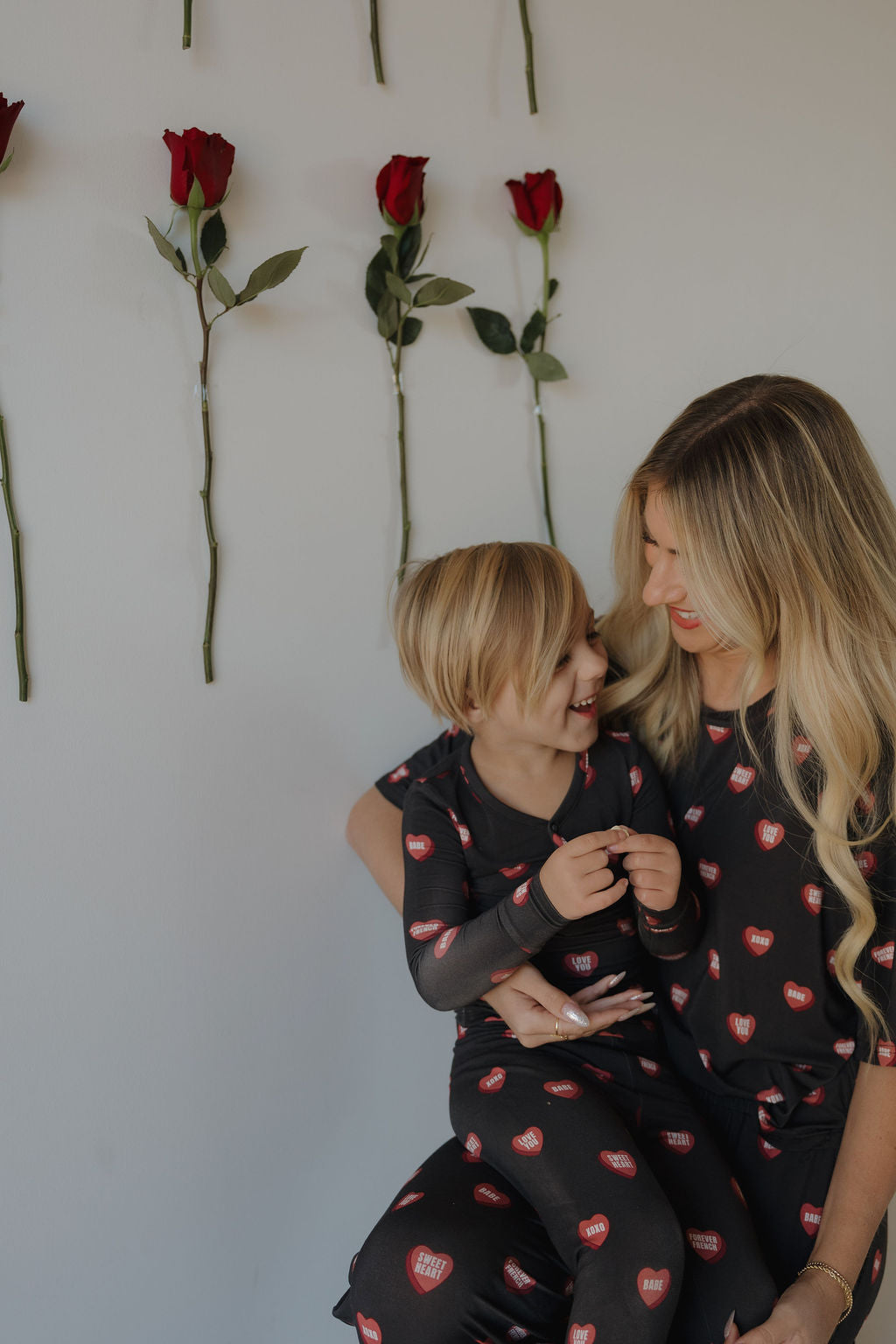 A woman and child with blonde hair smile at each other, both wearing matching forever french baby Love Notes Charcoal bamboo lounge sets. Red roses are attached vertically to the white wall behind them.