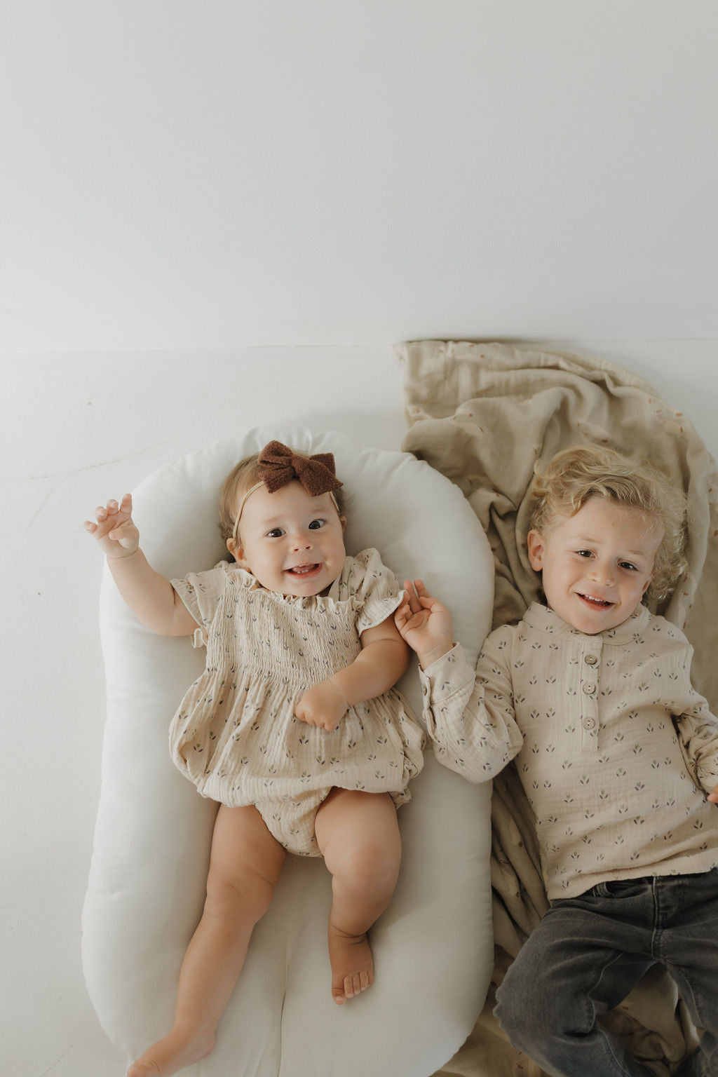 A baby girl in a brown bow and muslin cotton outfit lies on a white cushion, holding hands with a young boy wearing the Muslin Button Up | Flora from forever french baby, both smiling on a beige blanket and showcasing adorable kids' clothing.