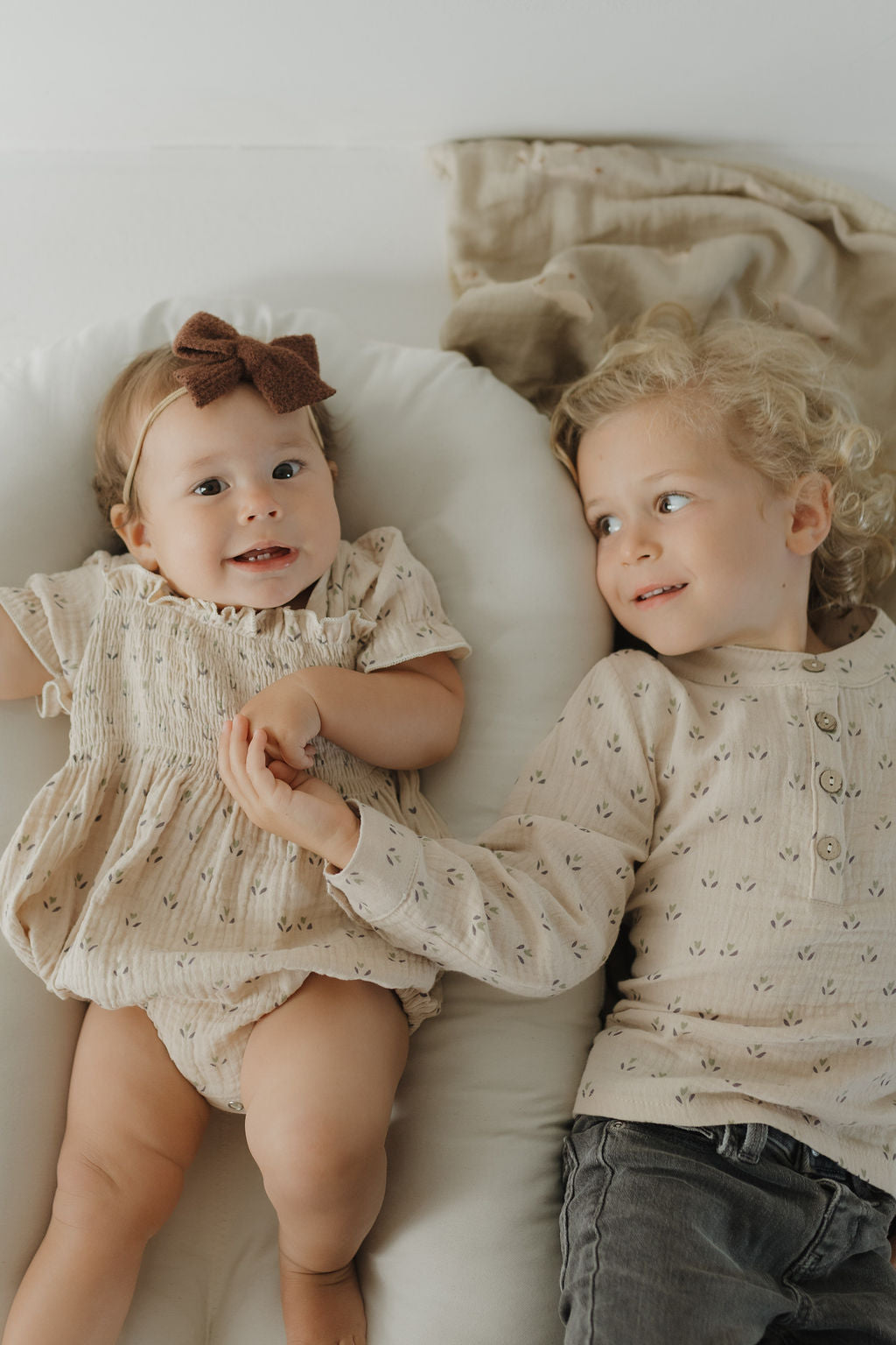 A baby girl in a brown bow headband and a toddler boy with curly hair lie on a bed, smiling and holding hands. Both wear the forever french baby Muslin Button Up | Flora—light, patterned muslin cotton for cozy kids’ style.