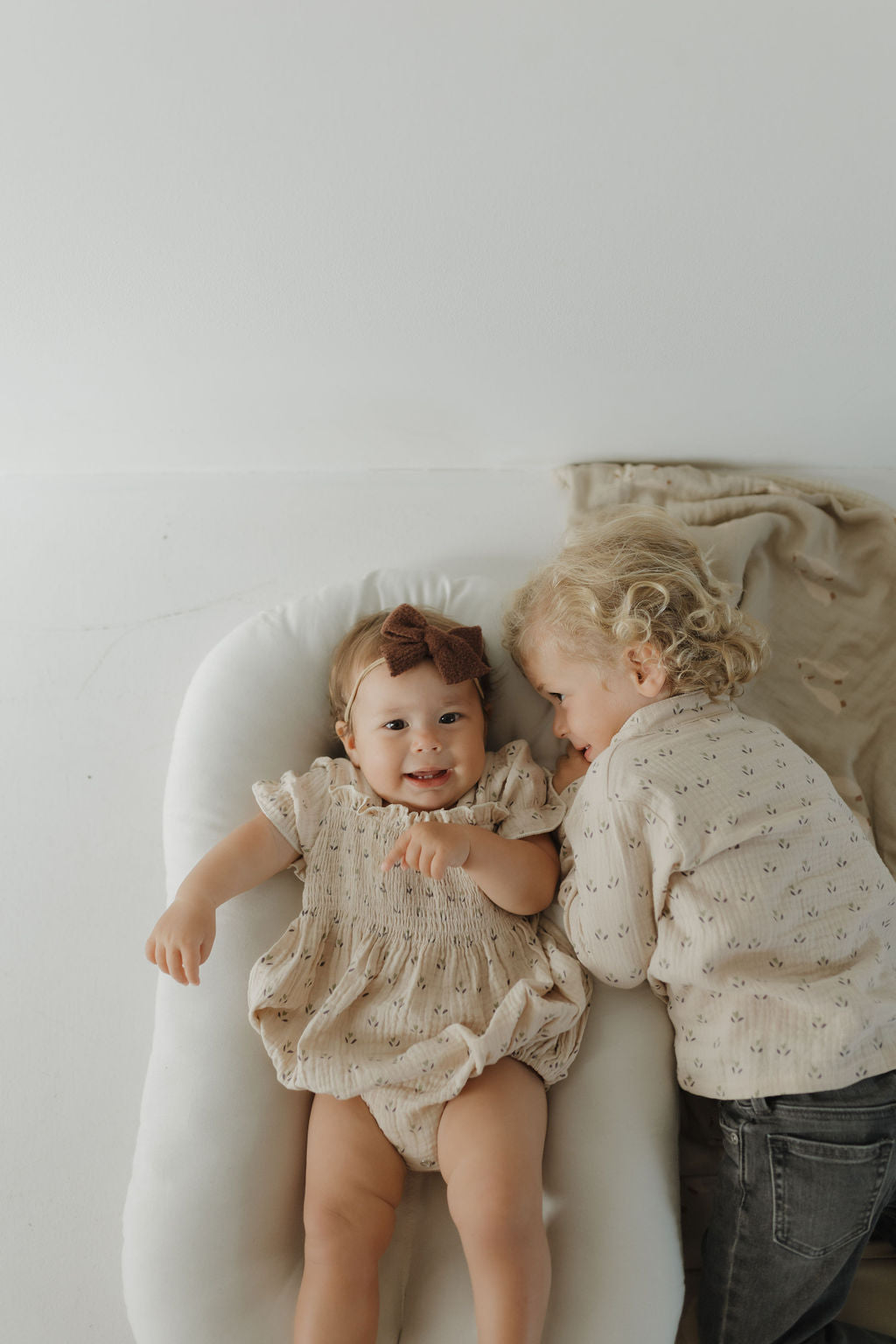 A smiling baby in the forever french baby Muslin Button Up | Flora lies on a white cushion, while an older child in matching kids' clothing rests beside them on a beige blanket.