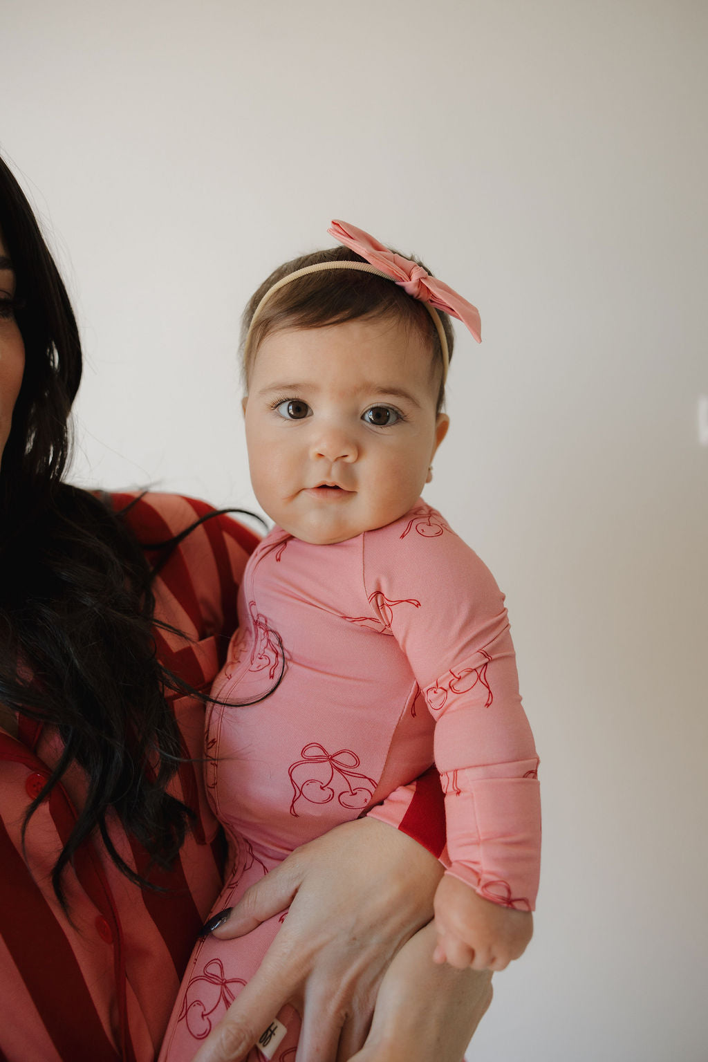 A baby in Minty Cherries Bamboo Zip Pajamas | Iconic Cherry—a pink outfit with red patterns and a matching pink bow headband—is held by an adult, gazing neutrally at the camera against a plain, light background.
