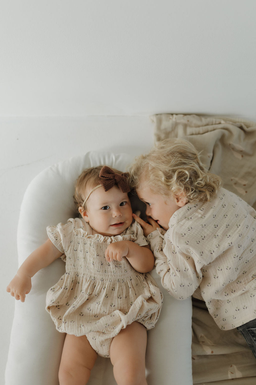 A baby in the forever french baby Muslin Button Up | Flora romper with a brown bow headband lies on a white cushion as a curly-haired child in a matching outfit leans in, smiling.