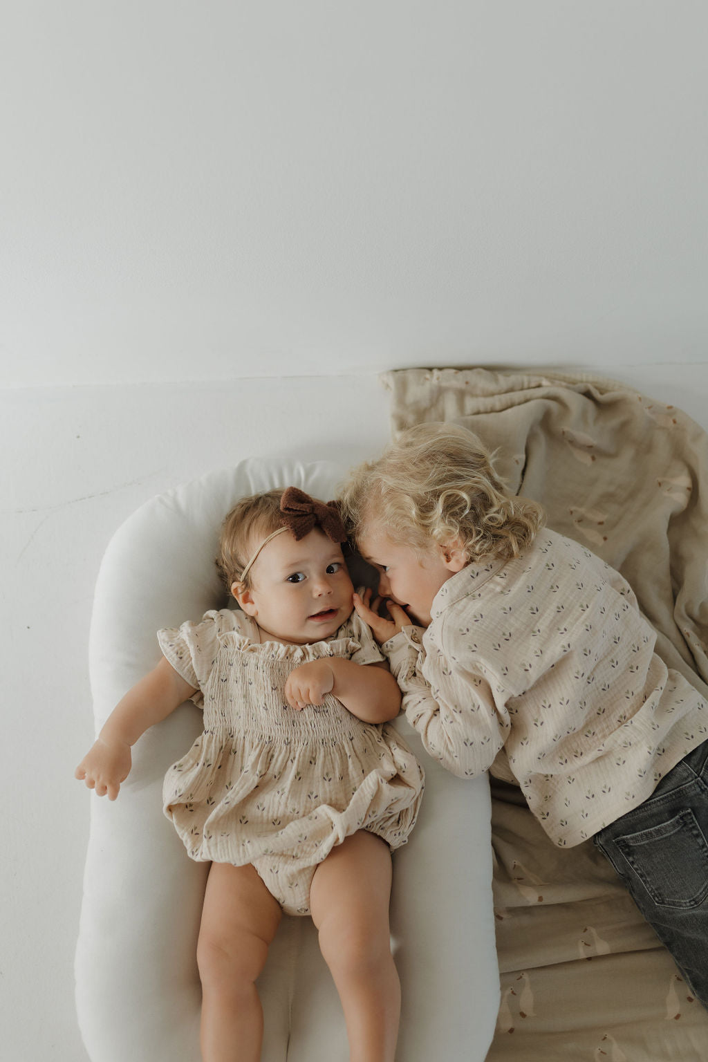A baby in the forever french baby Muslin Button Up | Flora romper lies on a white cushion, while a young child with curly blonde hair leans in, both dressed in light, patterned outfits for a matching moment.