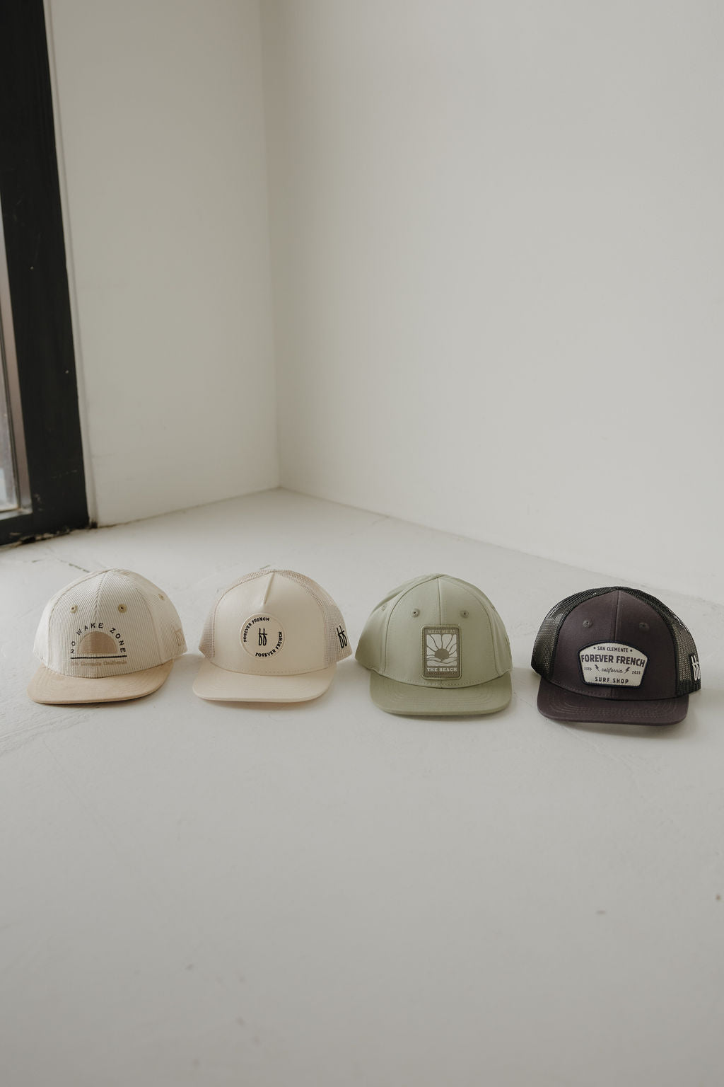 Four Snapback | ff Surf Shop caps by forever french baby—two beige, one olive green, one dark gray—are lined up on a white floor near a window. Each features a unique front design or patch for extra style.