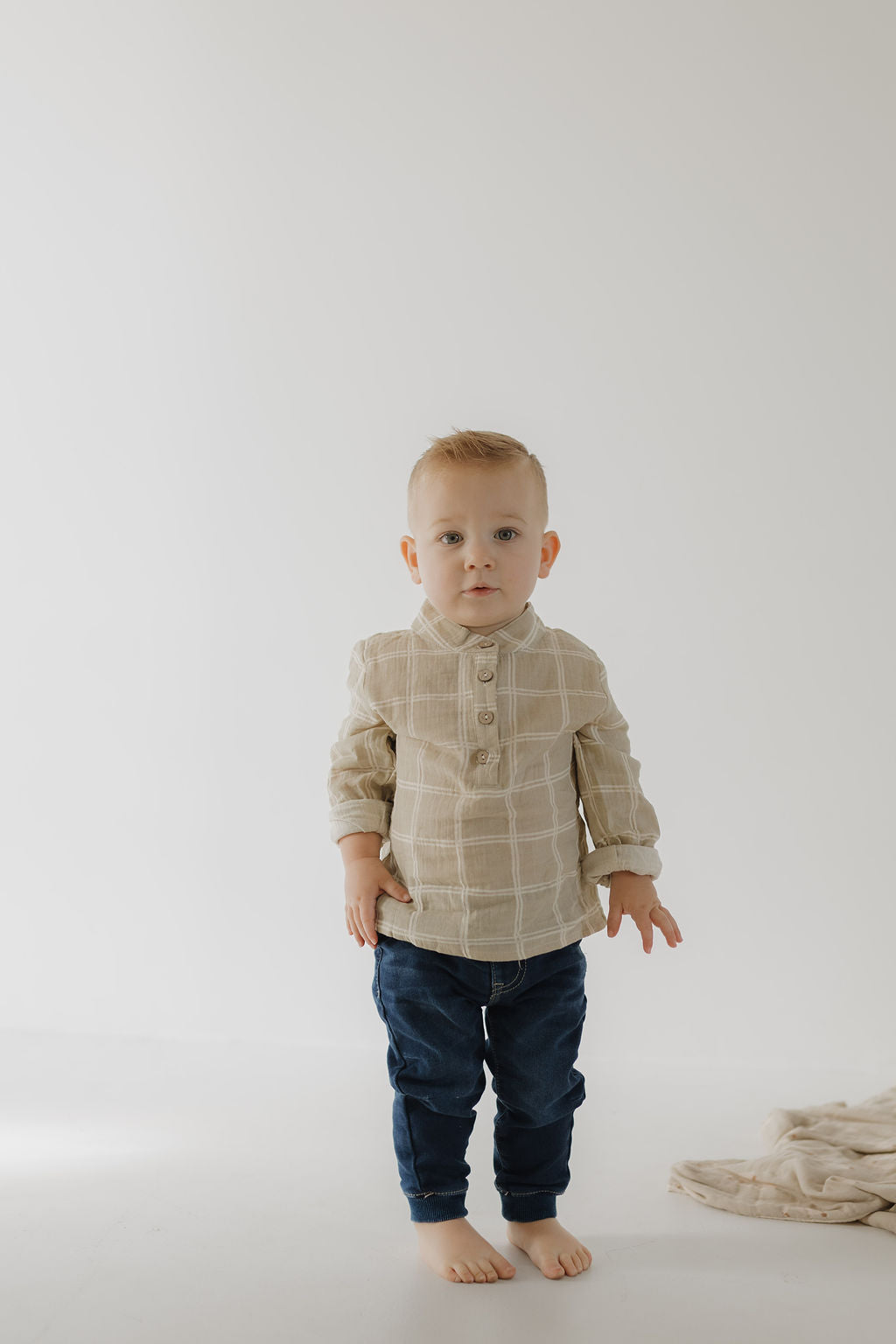 A young child with short blonde hair stands barefoot on a white floor, wearing the forever french baby Muslin Button Up | Linen Grid shirt and dark blue jeans. A matching soft muslin cotton blanket lies nearby against a plain white background.