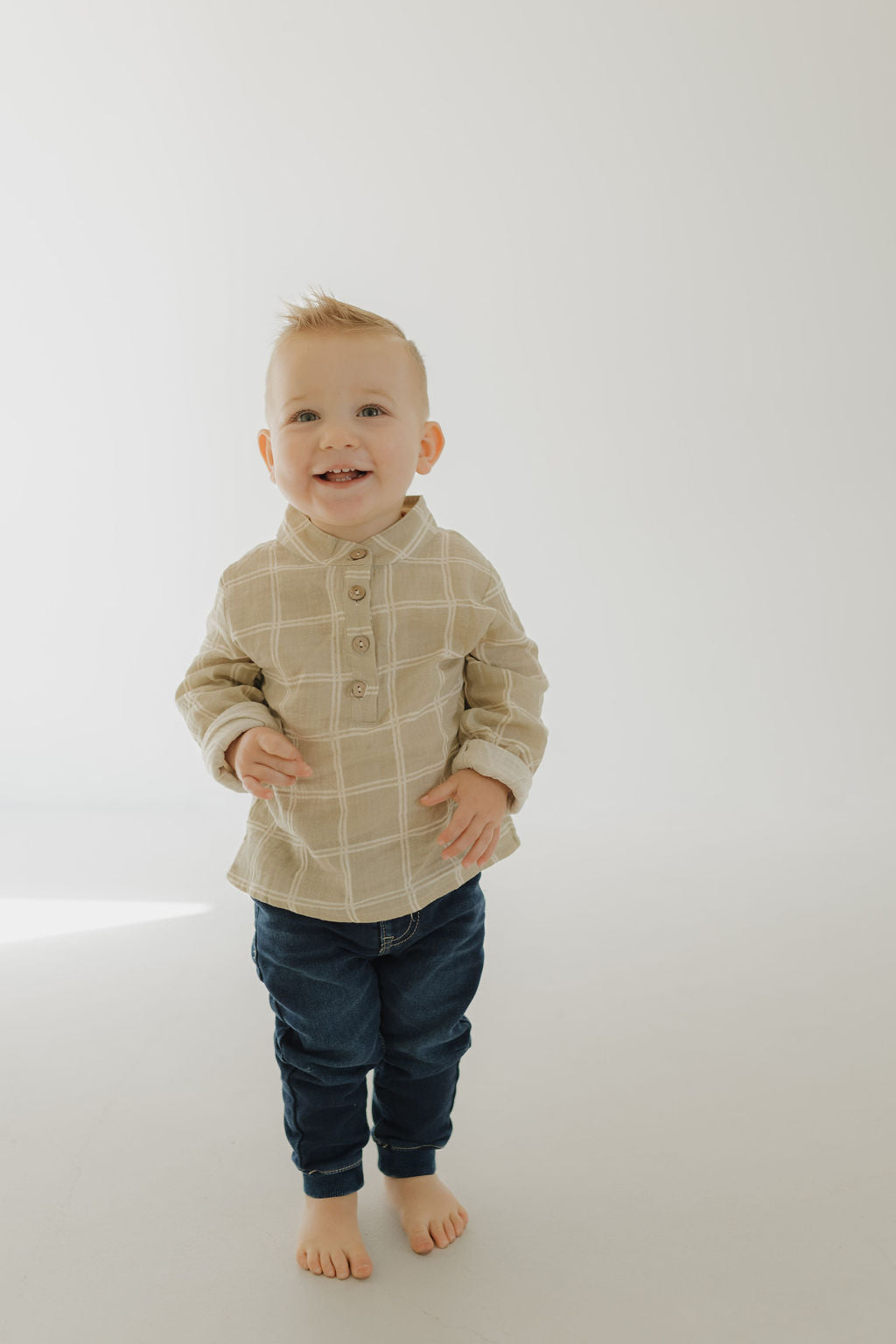 A smiling young child with short blonde hair stands barefoot on a light background, wearing the forever french baby Muslin Button Up | Linen Grid and dark blue jeans—perfect for matching styles with friends or siblings.
