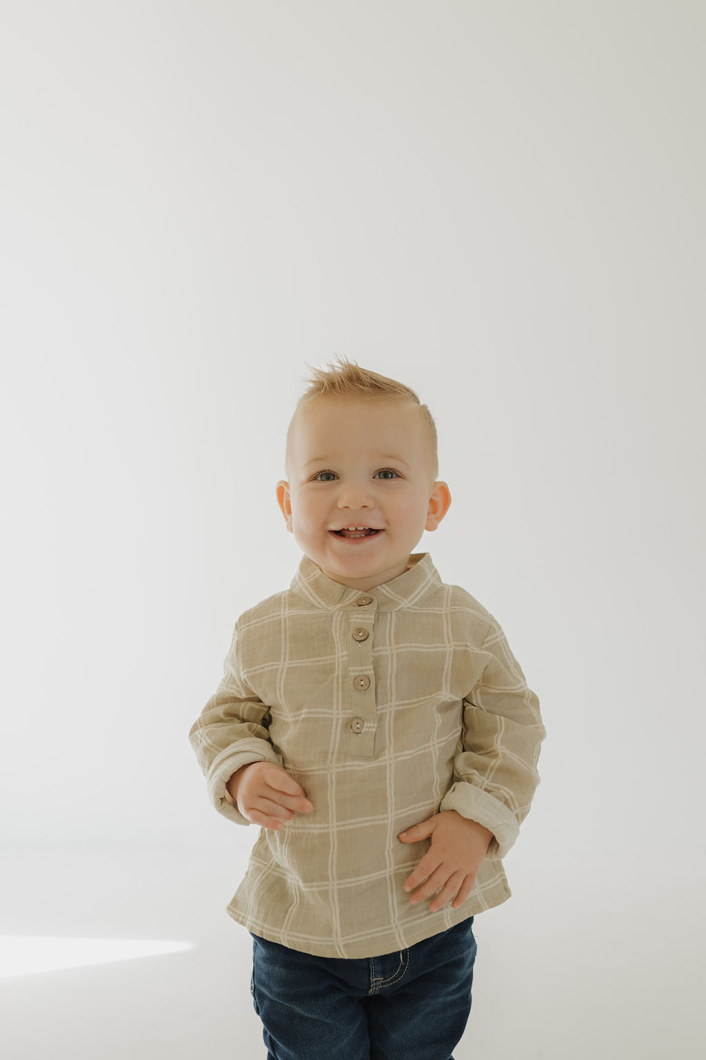 A smiling toddler with short blonde hair wears the forever french baby Muslin Button Up in Linen Grid, pairing soft, breathable muslin cotton style with blue jeans against a light background.