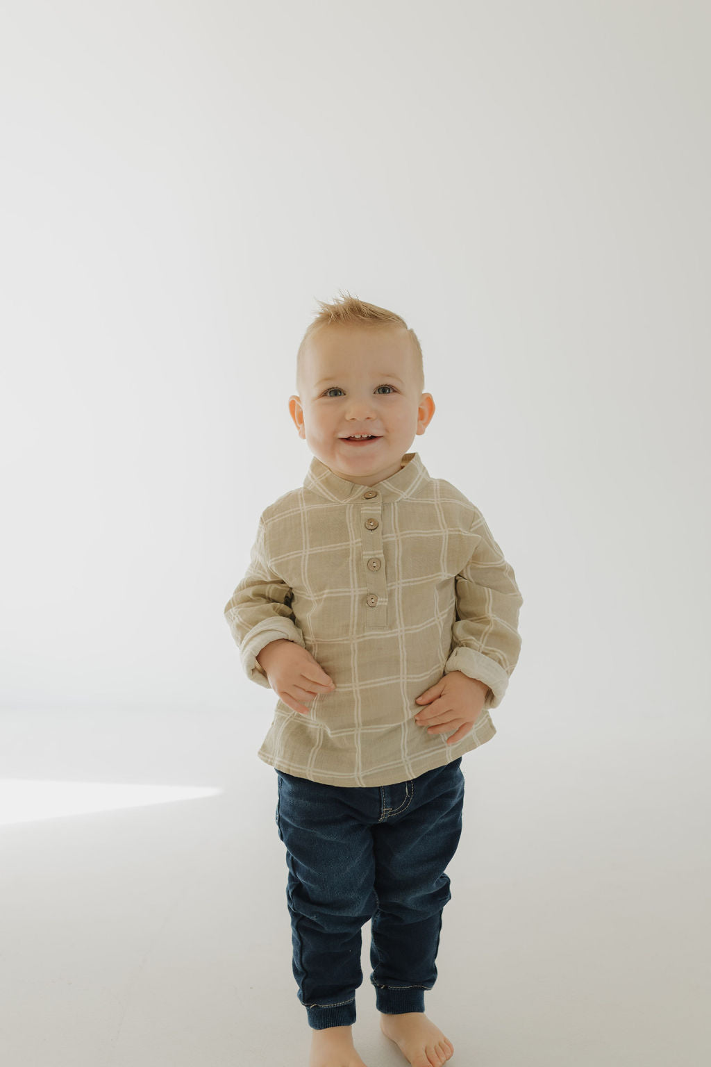 A young child with short blonde hair smiles while standing barefoot on a light floor, wearing the forever french baby Muslin Button Up in Linen Grid, paired with dark blue jeans against a plain light background.