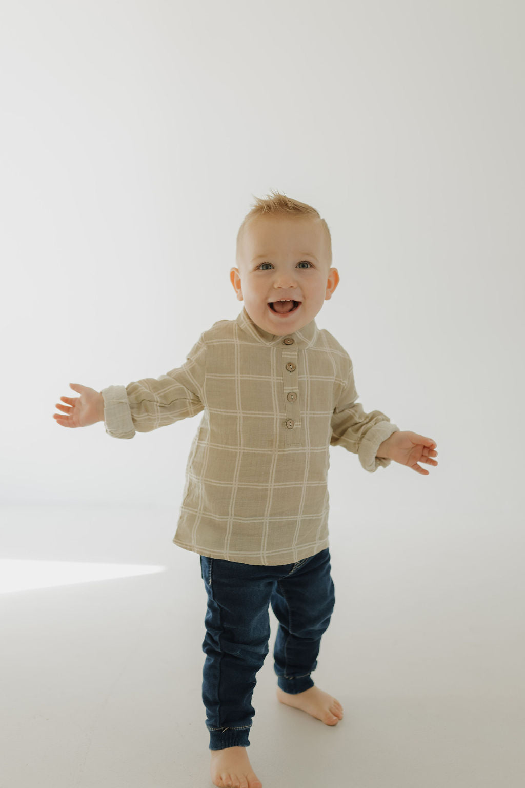 A smiling toddler stands barefoot on a light floor, wearing the forever french baby Muslin Button Up | Linen Grid, arms outstretched in a bright, minimal setting.