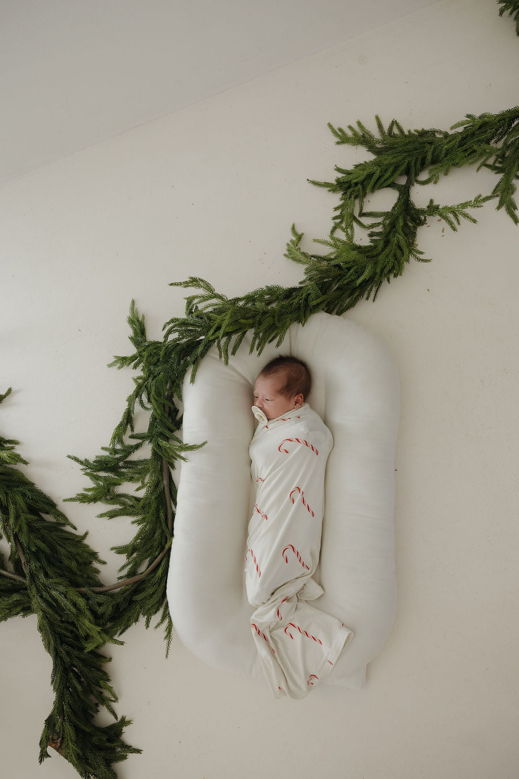 A baby wrapped in the forever french baby Bamboo Swaddle | Candy Cane (Preview) lies on a white cushion, surrounded by green pine garlands on a white floor.