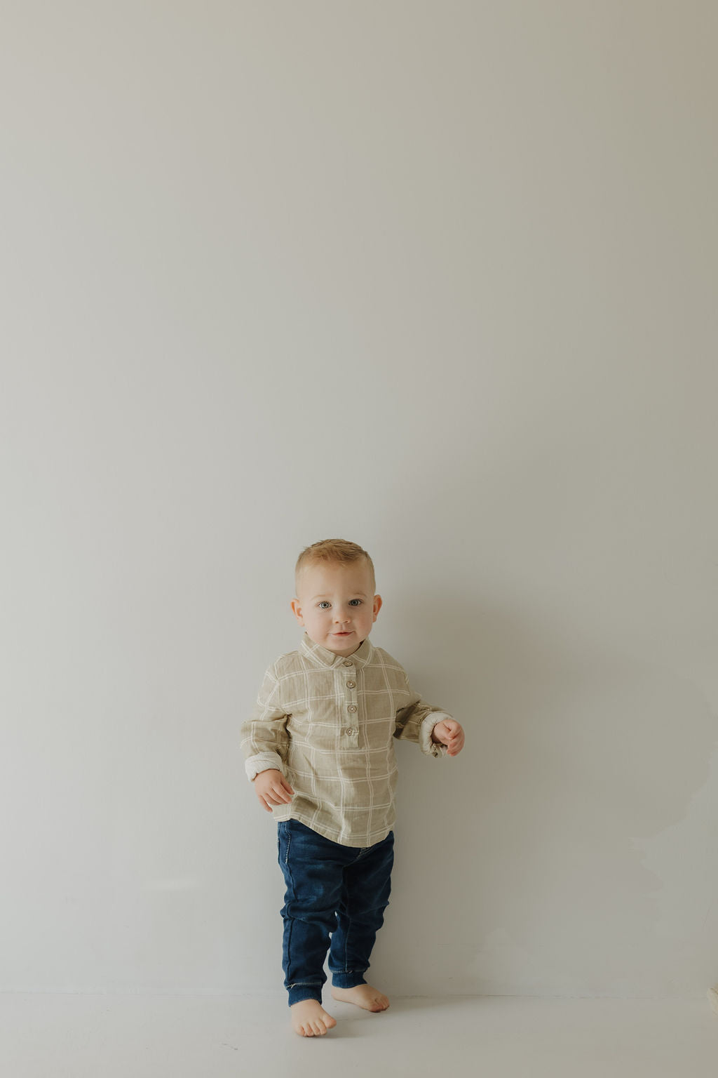 A young barefoot boy with short hair stands against a plain, light-colored wall, wearing the forever french baby Muslin Button Up | Linen Grid shirt and dark blue pants. He looks directly at the camera with a neutral expression in breathable, matching styles.