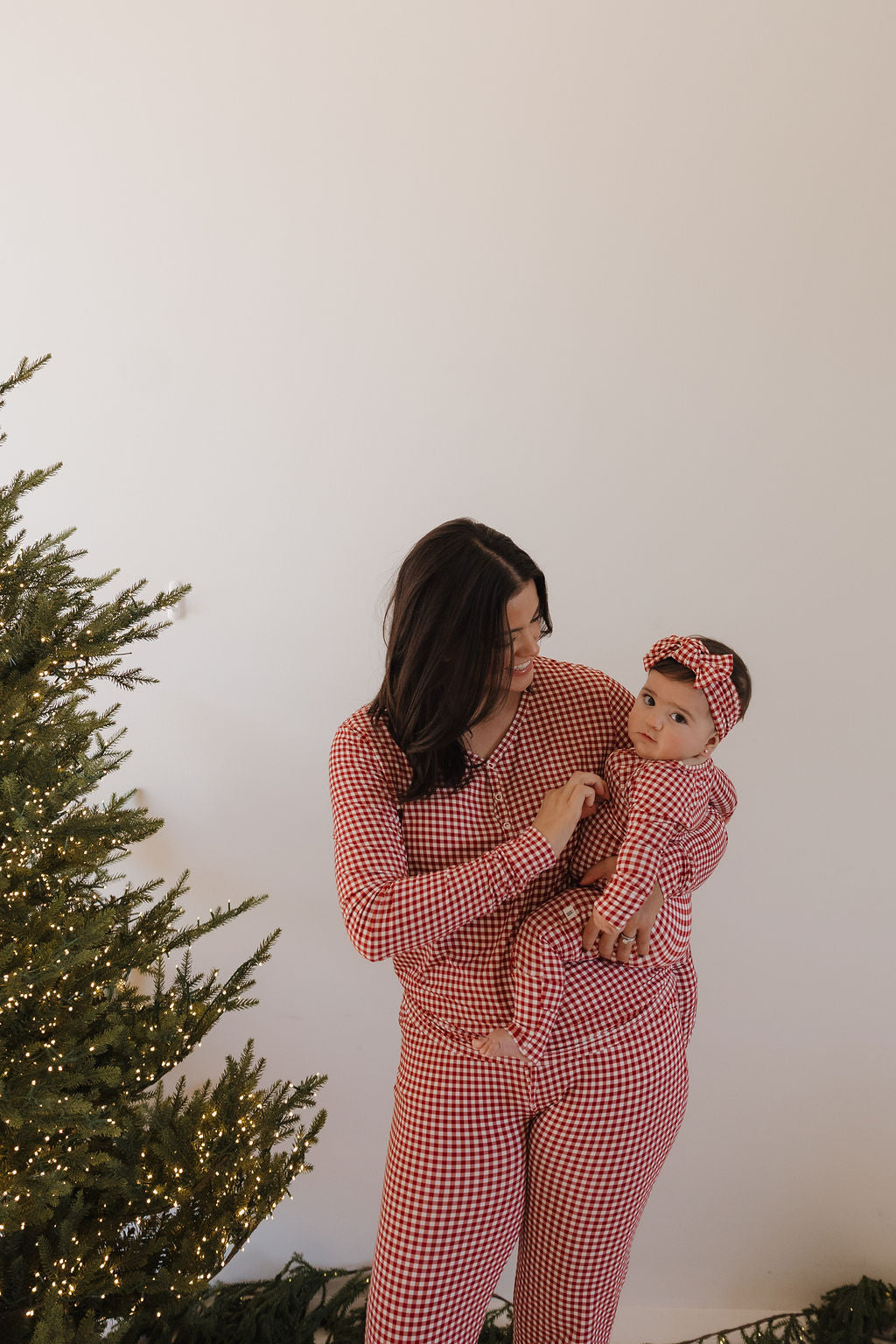 A woman wearing forever french baby's Women's Bamboo Pajamas in Red Gingham stands by a lit Christmas tree, holding a baby dressed in matching hypoallergenic pajamas, with a plain white wall as the backdrop.