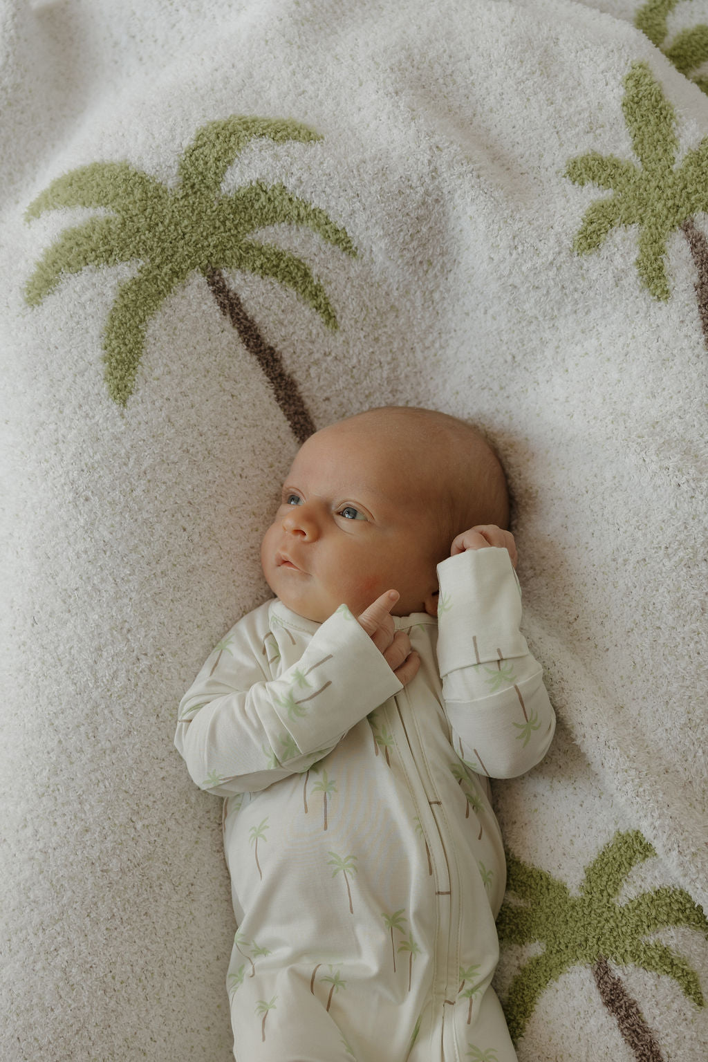 A baby in the forever french baby Bamboo Zip Pajamas | Palm, featuring a white fabric with green palm tree prints, lies on a soft white blanket adorned with green palms—comfortably dressed in breathable, gentle sleepwear ideal for delicate skin.