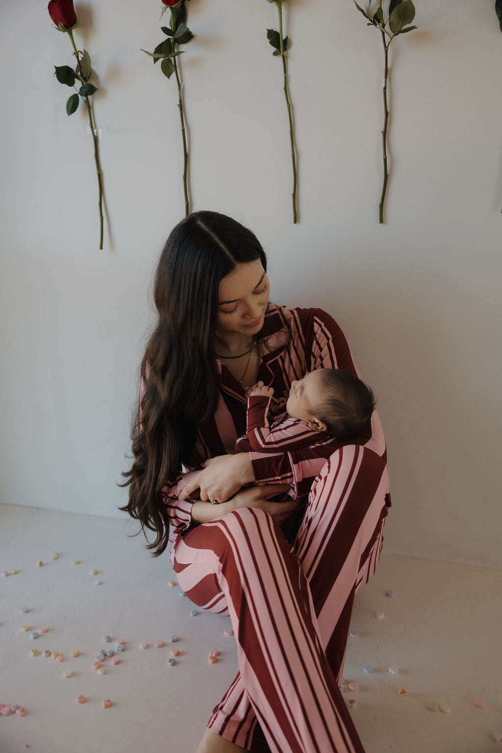 A woman in the forever french baby Women's Bamboo Button Up Lounge Set | Sweetheart Stripes sits on the floor holding a baby in matching pajamas, both gazing at each other. Three red roses and scattered petals decorate the scene behind them.