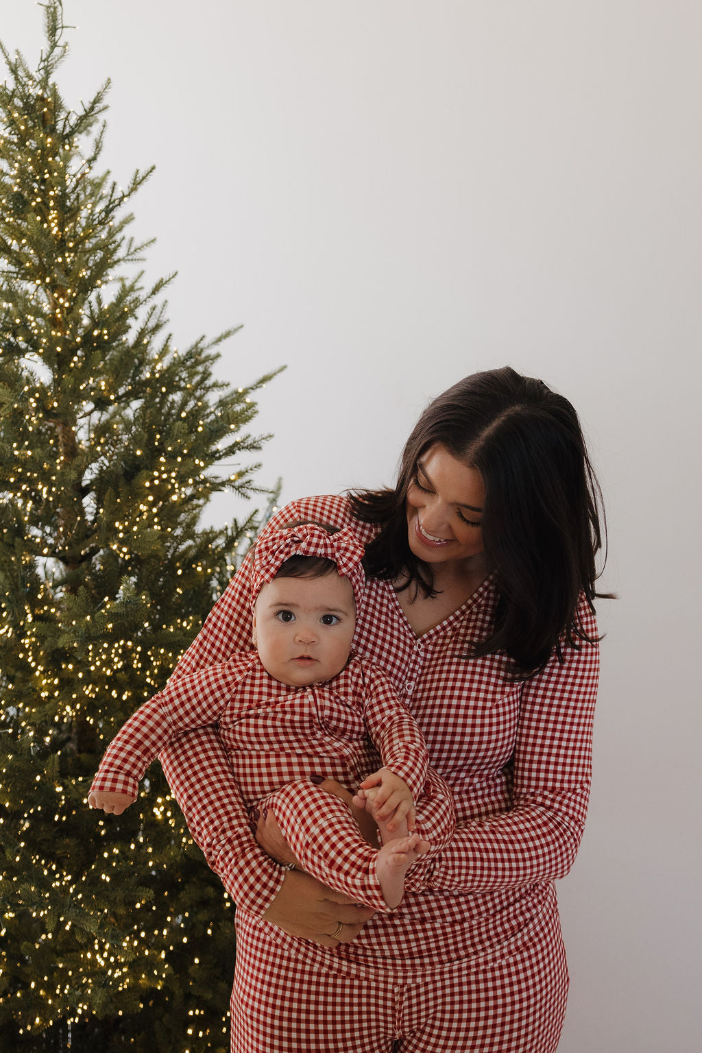 A woman and a baby wear matching forever french baby Women's Bamboo Pajamas in Red Gingham as they stand by a Christmas tree with white lights; the woman smiles at the baby, who looks at the camera.