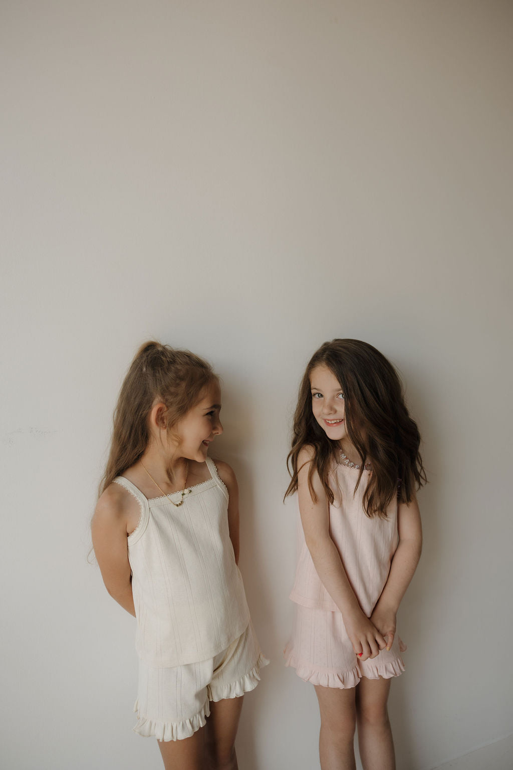 Two young girls stand against a plain light-colored wall, smiling and looking at each other. Both are wearing the Girl’s Ruffle Short Set in the Swan print from forever french baby—a charming summer outfit featuring light-colored sleeveless tops and matching ruffle shorts, perfect for warm days.