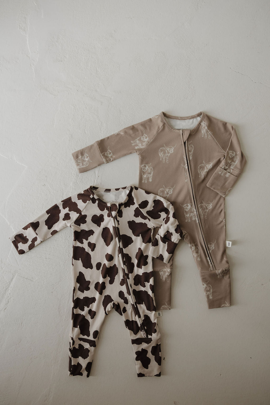 Two forever french baby Bamboo Zip Pajamas | Moo! are shown on a light textured surface—one in light brown with white animal line drawings, front zipper; the other in a brown and white cow print, diagonal zipper—crafted from hypoallergenic fabric.