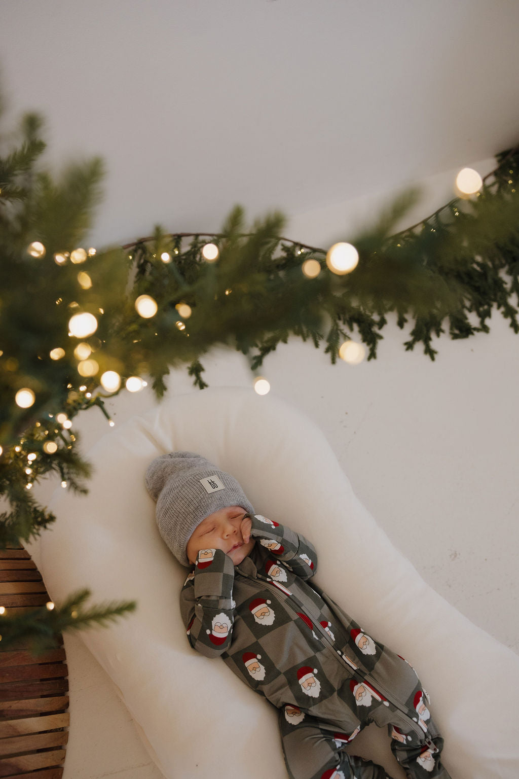 A baby in forever french baby Bamboo Zip Pajamas | Santa Checkerboard and a gray beanie sleeps on a white cushion near a decorated Christmas tree with string lights and festive greenery in the foreground.