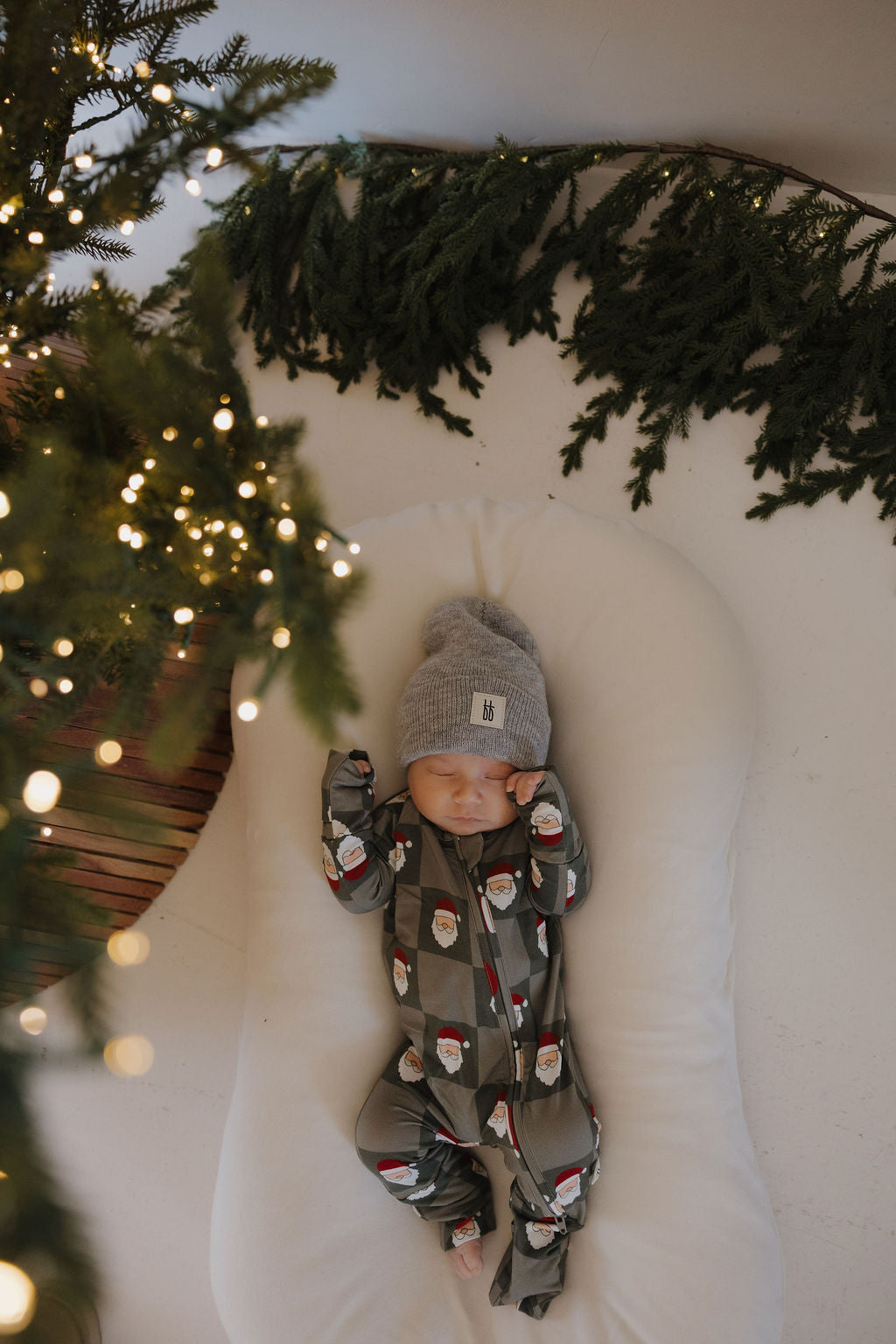 A baby in forever french baby Bamboo Zip Pajamas | Santa Checkerboard and a gray hat lies on a white cushion beside a Christmas tree and green garland, showcasing festive and adorable holiday sleepwear.