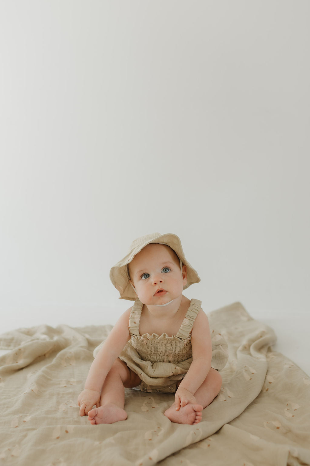 A baby in a beige outfit and sunhat sits barefoot on the forever french baby Muslin Quilt | Silly Goose against a light background, gazing up with curiosity—an ideal touch for any cozy nursery.