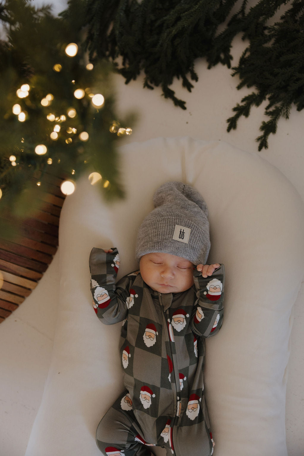 A baby in forever french baby's Bamboo Zip Pajamas | Santa Checkerboard and a gray beanie sleeps peacefully on a white cushion near a glowing Christmas tree, perfectly dressed for the festive season.