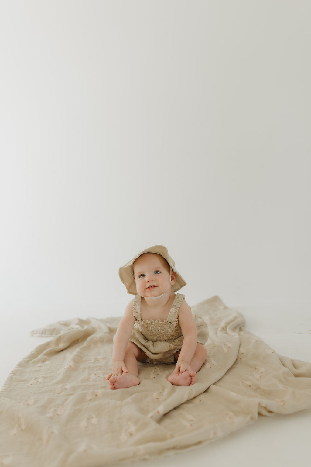 A baby in a beige outfit and sun hat sits on the Muslin Quilt | Silly Goose by forever french baby, atop a light backdrop—an adorable choice for a cozy nursery.