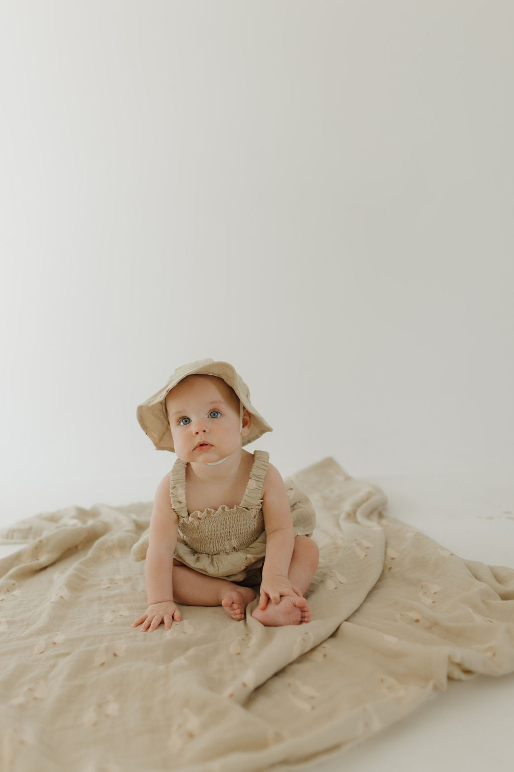 A baby in a beige romper and sun hat sits on the forever french baby Muslin Quilt | Silly Goose, set against a light background—an adorable addition to any nursery.