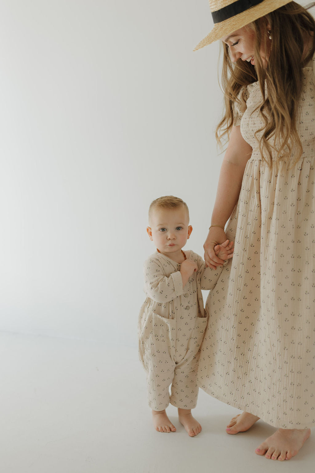 A baby in light beige holds hands with an adult wearing the forever french baby Women's Short Sleeve Muslin Dress | Flora—both barefoot on a white background, creating a perfect mom-and-me matching look.