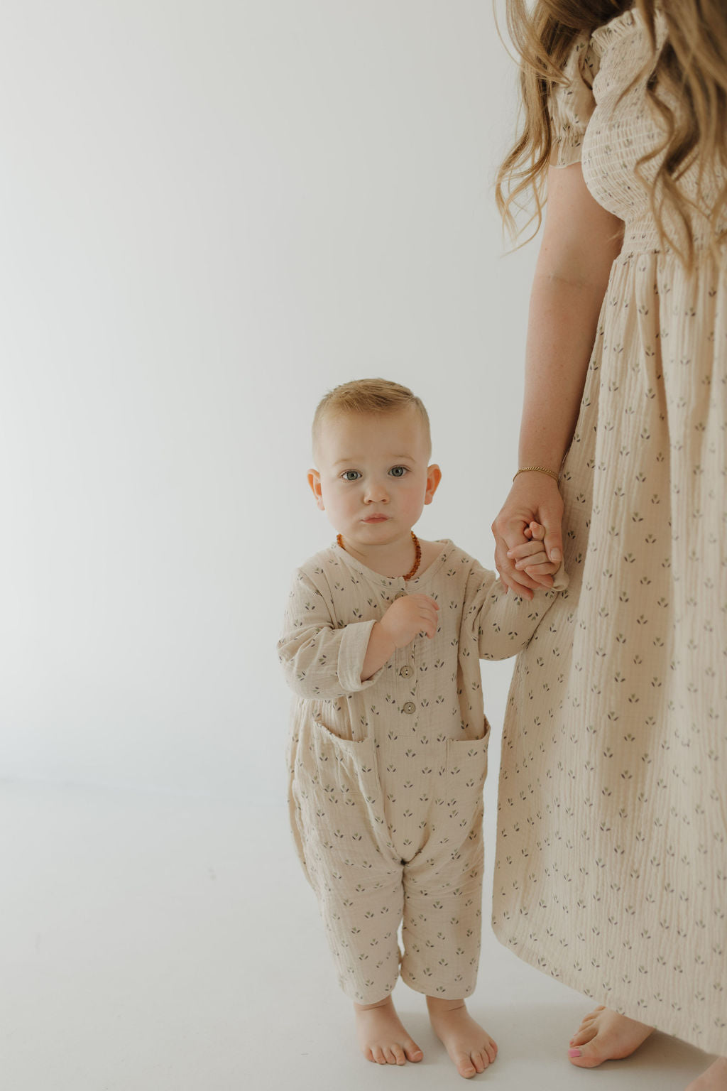 A young child in beige stands barefoot, holding the hand of an adult woman wearing the forever french baby Women's Short Sleeve Muslin Dress | Flora. Both are partially visible against a plain, light background.