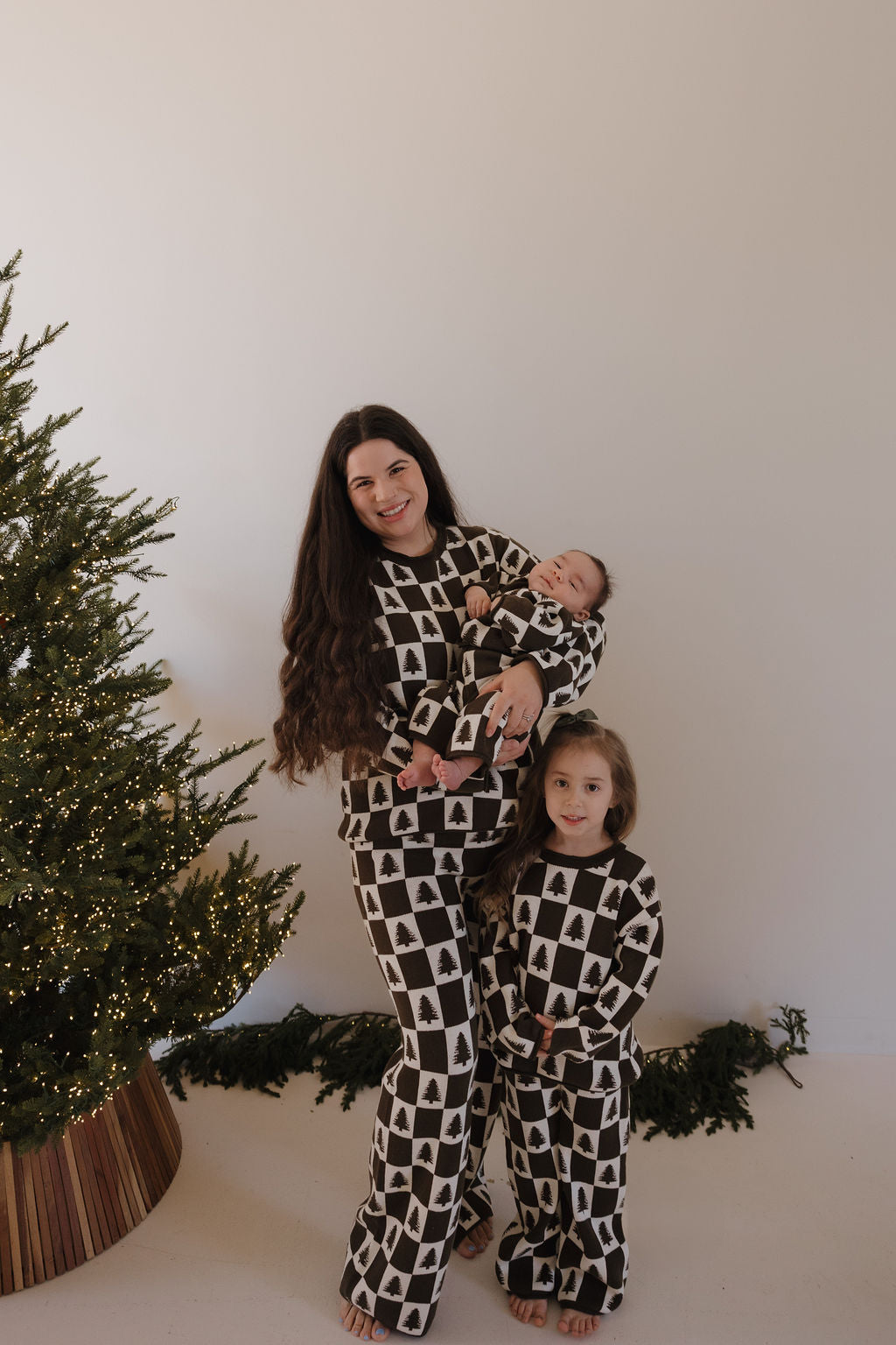 A woman, baby, and young child in matching forever french baby Women's Knit Pant Set | Christmas Tree Checkerboard pajamas pose together by a lit Christmas tree in a minimal, festive room.