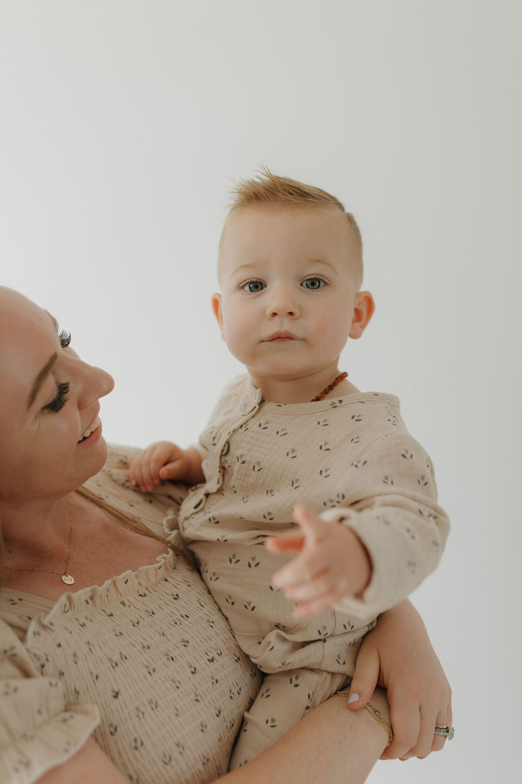 A woman smiles while holding a baby dressed in the forever french baby Women's Short Sleeve Muslin Dress | Flora. The baby reaches toward the camera, their sweet bond highlighted by a light, minimalistic background.