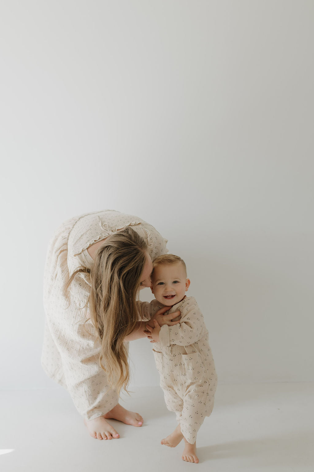 A woman wearing the Women's Short Sleeve Muslin Dress | Flora by forever french baby bends to kiss a smiling baby. Both are barefoot in light, patterned outfits, standing against a plain, light background.