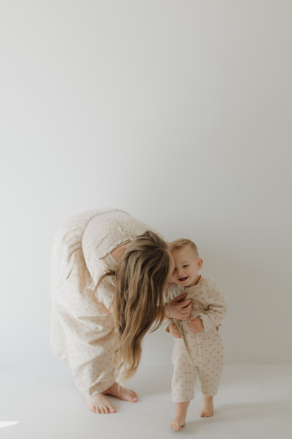 A woman in the "Women's Short Sleeve Muslin Dress | Flora" by forever french baby bends to embrace a smiling toddler; both are barefoot, wearing light patterned muslin dresses against a plain, light background.